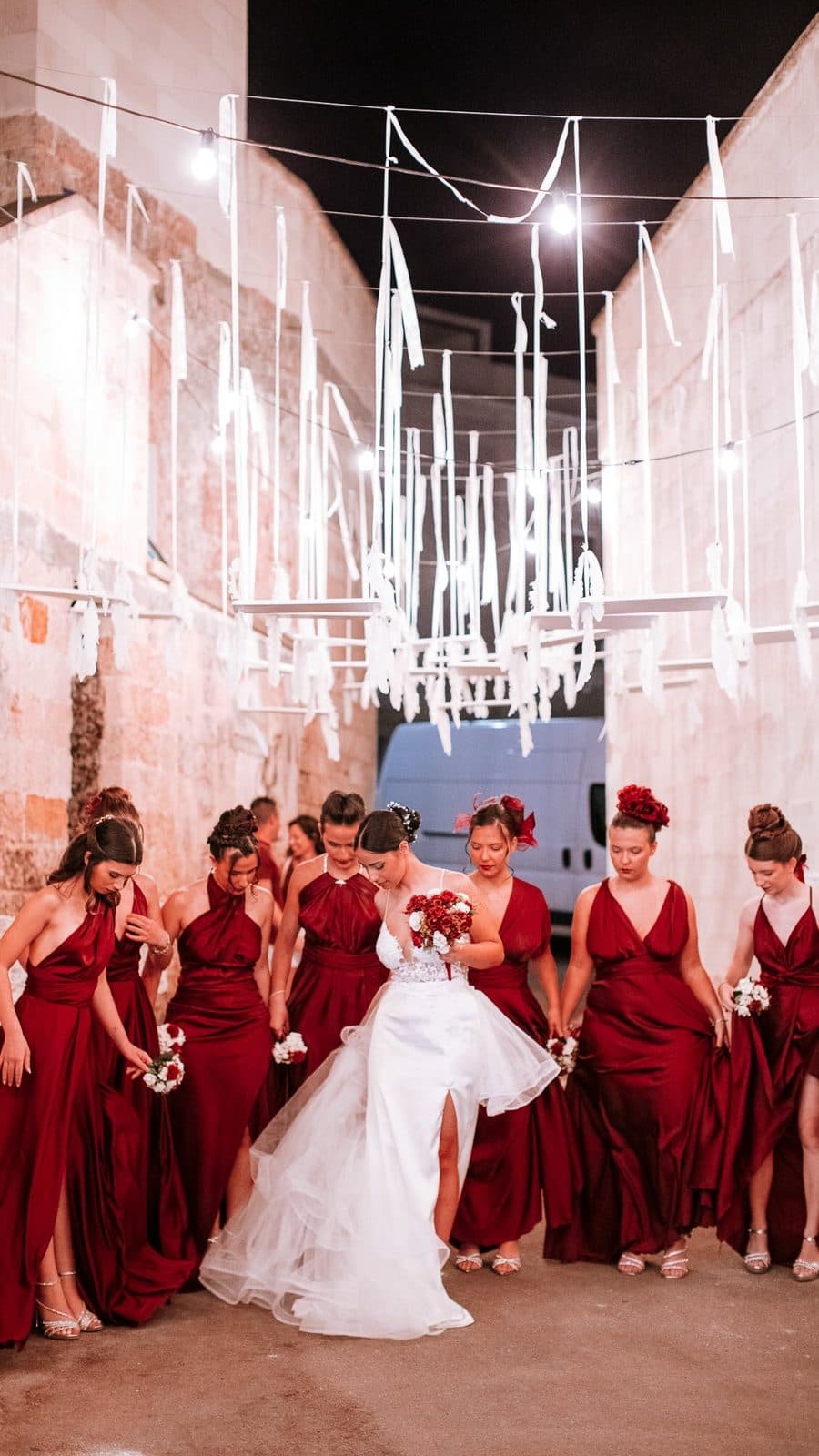 A bride and her bridesmaids in red under an installation of white ribbons in a stone alley