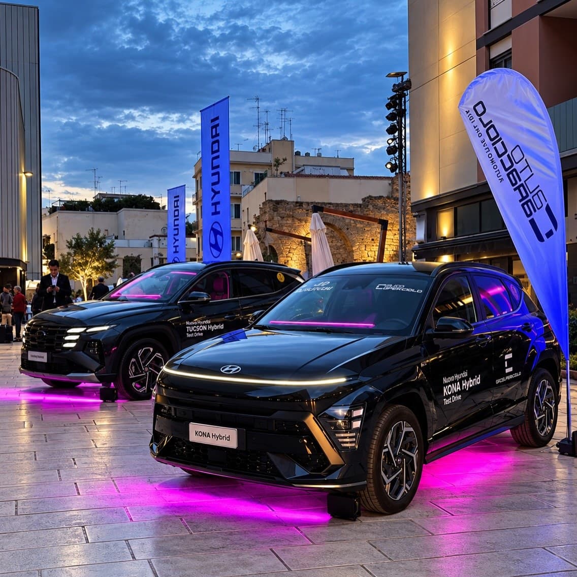 Cars lit from below with magenta LED bars at a launch in a town square, at dusk